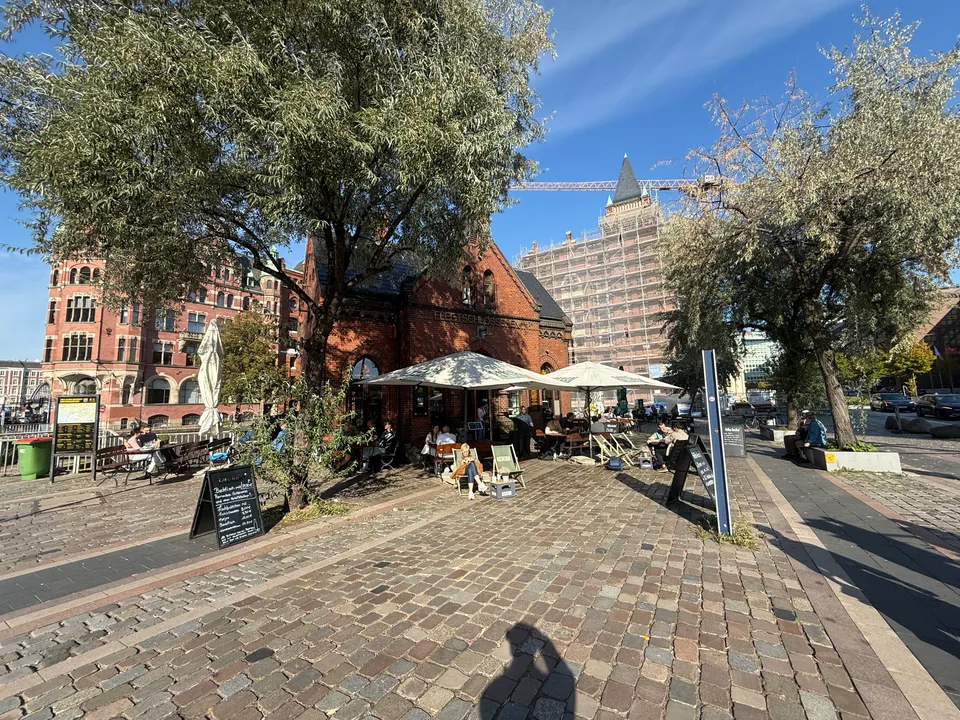 Kopfsteinpflasterplatz mit Außengastronomie, Sonnenschirmen und Liegestühlen vor kleinem Backsteinhaus, Bäume und Speicherstadtgebäude im Hintergrund