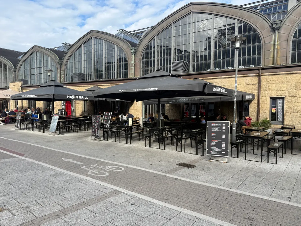 Außenterrasse mit schwarzen Sonnenschirmen und Tischen vor den Rundbogenfenstern einer großen Bahnhofshalle, davor ein Radweg