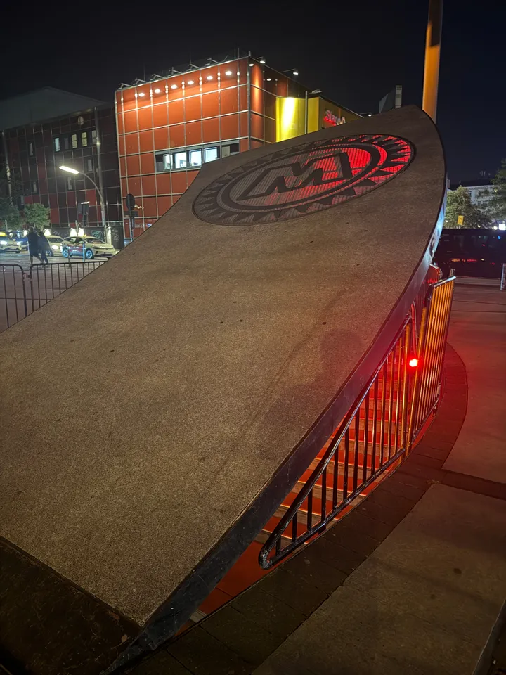 Geneigte graue Deckelplatte mit rot leuchtendem M-Logo, daneben beleuchtete Treppe, dahinter rotes Gebäude