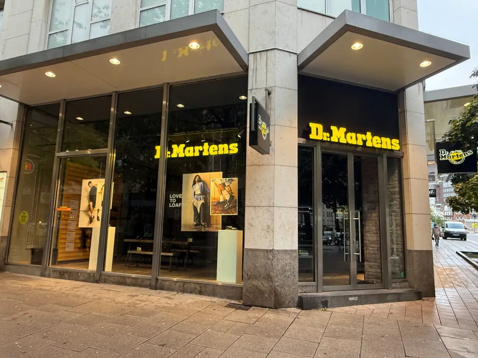 Eckgeschäft von Dr. Martens mit gelber Leuchtschrift, Glasschaufenstern und Natursteinfassade an einer gepflasterten Einkaufsstraße