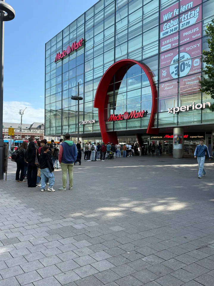 Glasfassade eines MediaMarkt mit rotem Bogen über dem Eingang, davor eine lange Schlange wartender Menschen auf gepflastertem Platz