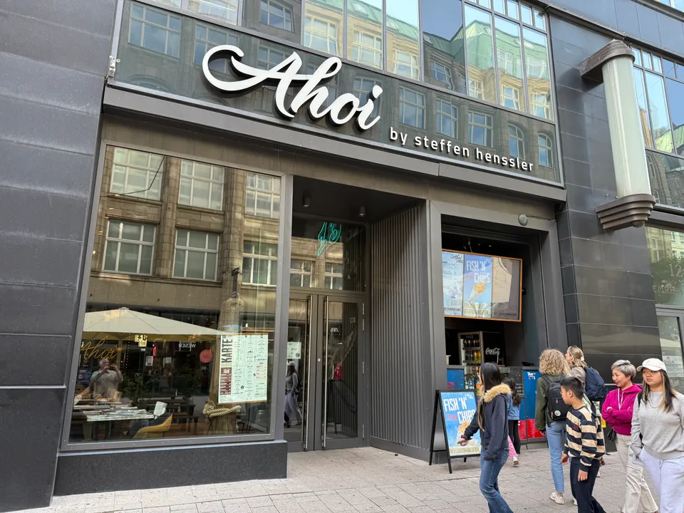 Restaurantfassade „Ahoi by Steffen Henssler“ mit Glasfront, Fish-'n'-Chips-Aufsteller und Passanten in einer Hamburger Einkaufsstraße