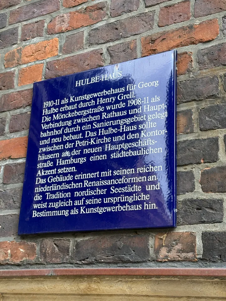 Blaue Emailtafel „Hulbe-Haus“ mit Text zur Baugeschichte an einer Backsteinfassade