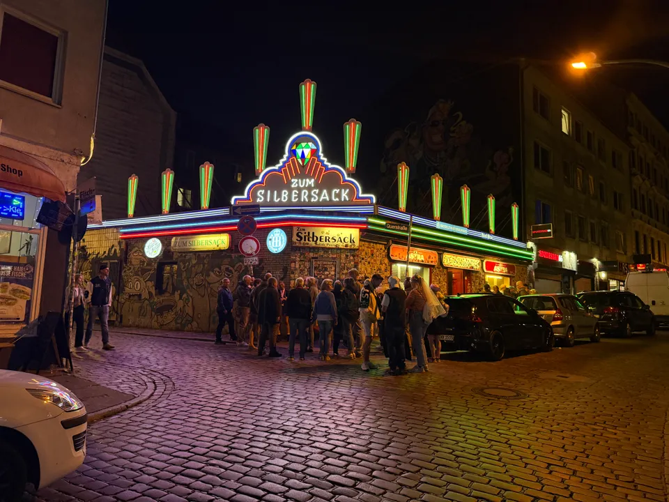Eckkneipe mit bunter Neon-Leuchtreklame bei Nacht, davor eine wartende Menschengruppe auf Kopfsteinpflaster