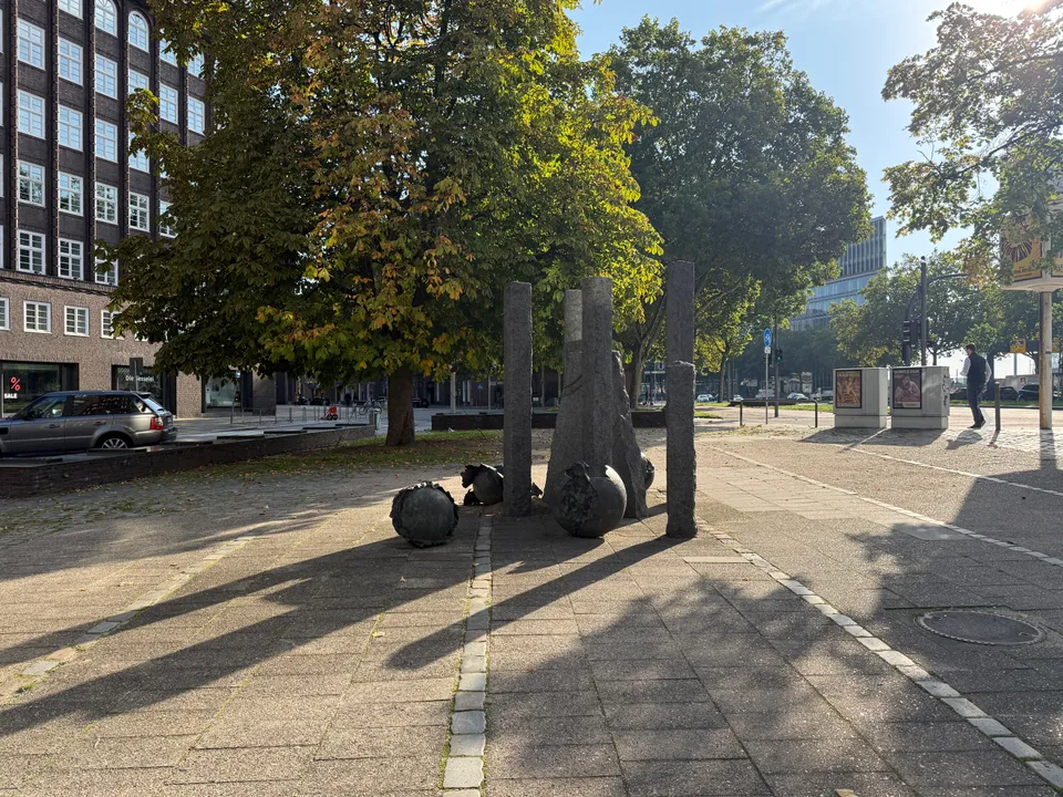 Granitstelen und aufgebrochene Bronzekugeln auf gepflastertem Platz vor Kastanienbaum und dunklem Klinker-Bürogebäude