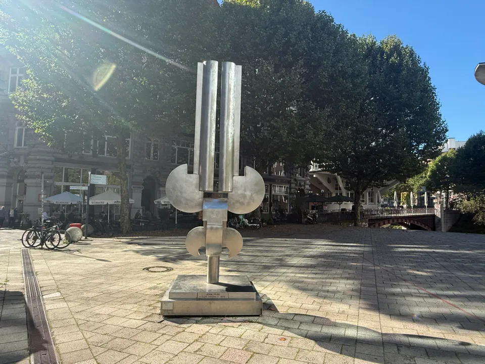 Edelstahlskulptur aus senkrechten Rohren und halbrunden Scheiben auf gepflastertem Platz, dahinter Bäume, Altbau und Fleetbrücke