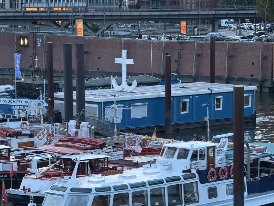 Blauer schwimmender Gemeindebau mit weißem Ankerkreuz zwischen Barkassen, dahinter Kaimauer und Hochbahnbrücke