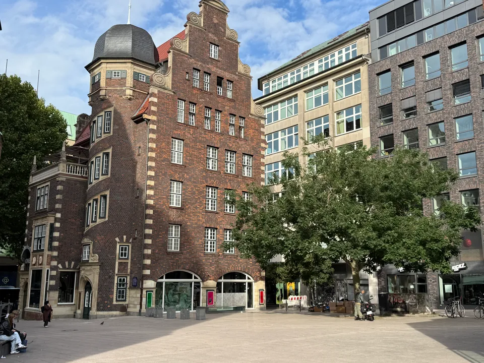 Backsteingebäude mit Treppengiebel und Rundturm an einem Platz, daneben moderne Geschäftshäuser und ein Laubbaum