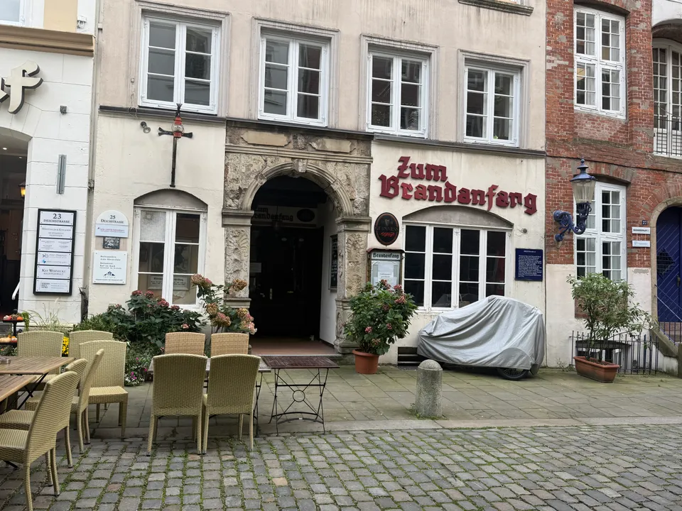 Fassade der Gaststätte Zum Brandanfang mit Sandsteinportal, roter Frakturschrift, Hortensien und leeren Rattanstühlen auf Kopfsteinpflaster