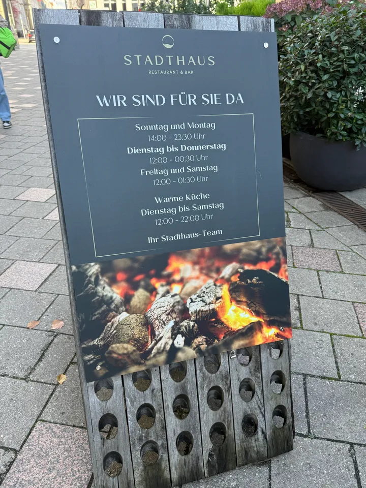 Hölzerner Aufsteller mit Öffnungszeiten des Stadthaus Restaurant & Bar und Foto glühender Holzkohle auf Pflaster