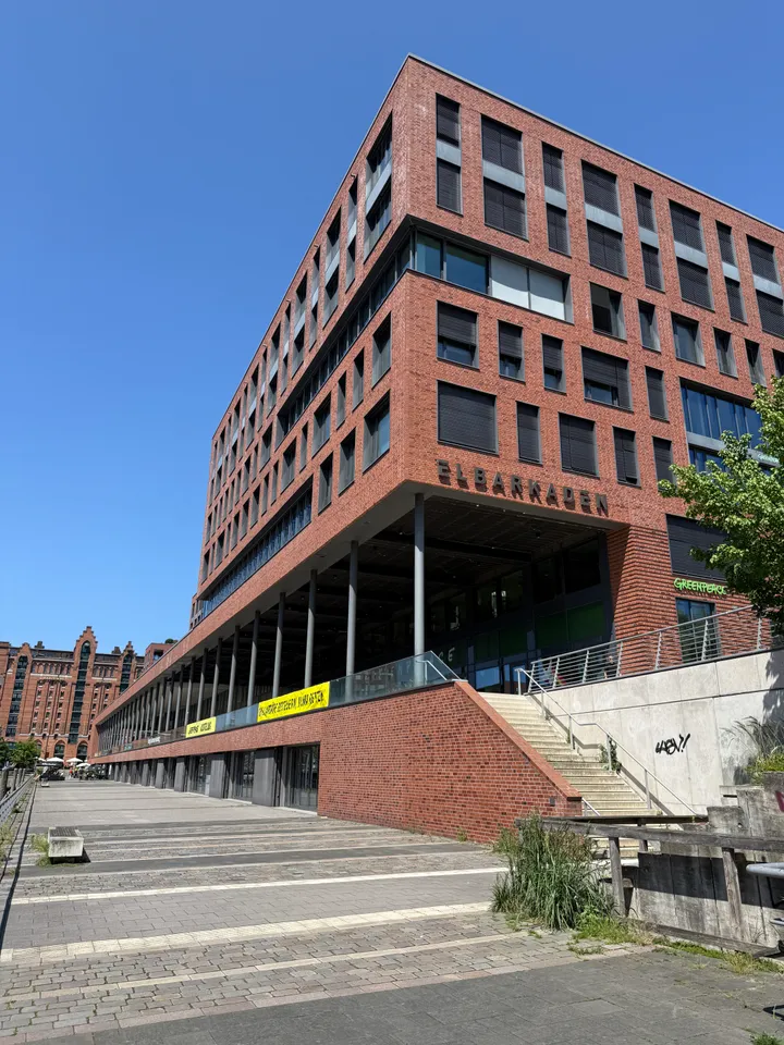 Rotes Klinkergebäude mit Schriftzug Elbarkaden, Säulengang im Erdgeschoss und gepflasterter Promenade unter blauem Himmel