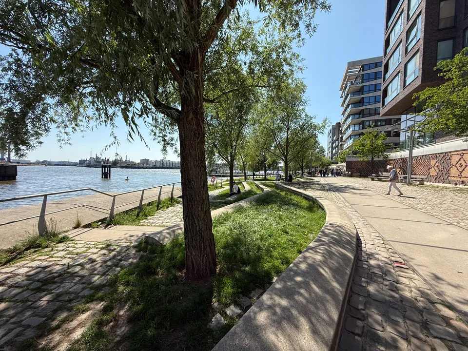 Uferpromenade mit Weiden, geschwungenen Betonsitzstufen, Rasen und modernen Wohnhäusern am Wasser bei Sonnenschein