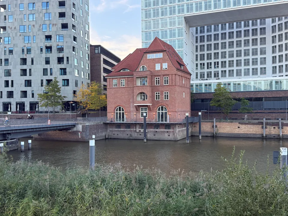 Rotes Backsteinhaus auf Kaimauer am Hafenbecken, links Brücke und Wohnhochhaus, rechts großes Glasgebäude