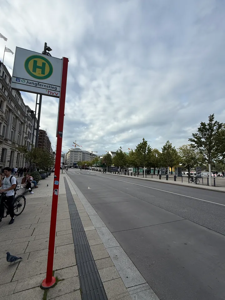 Haltestellenschild Jungfernstieg an roter Stange, daneben breite Straße und Baumreihe an der Alsterpromenade