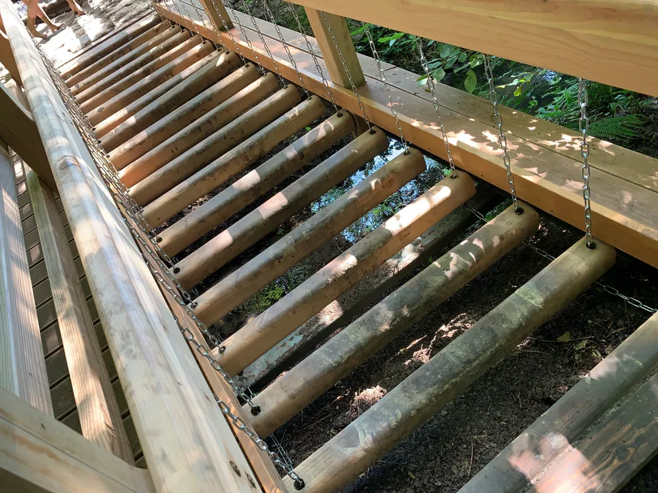 Hängebrücke aus an Ketten befestigten Rundhölzern über einem flachen Wassergraben, daneben ein hölzerner Handlauf