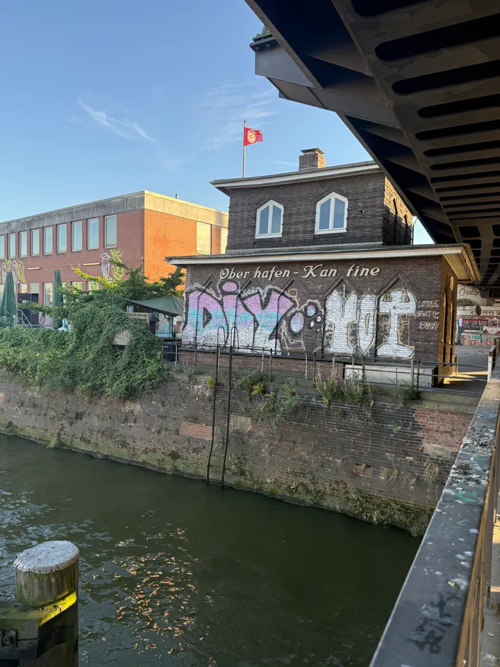 Backsteinhaus der Oberhafen-Kantine mit Graffiti an einer Kaimauer, darüber eine Stahlbrücke, davor ein Kanal