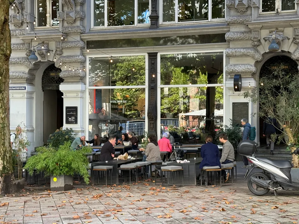 Außenterrasse einer Bar vor gründerzeitlicher Stuckfassade mit großen Schaufenstern, Gästen an Tischen und Herbstlaub