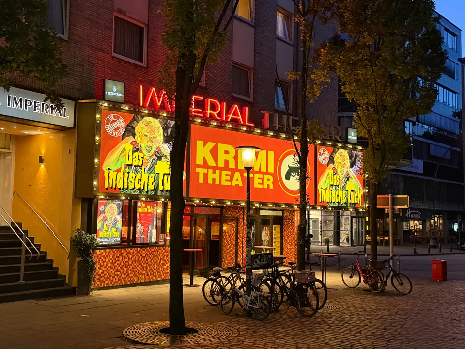 Beleuchtete Fassade des Imperial Theaters mit Krimi-Theater-Werbung, davor Fahrräder und Bäume in der Abenddämmerung