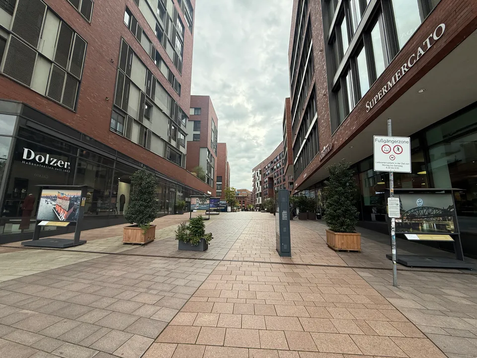 Leere gepflasterte Fußgängerzone zwischen Klinkerbauten mit Pflanzkübeln, Fototafeln und den Geschäften Dolzer und Supermercato