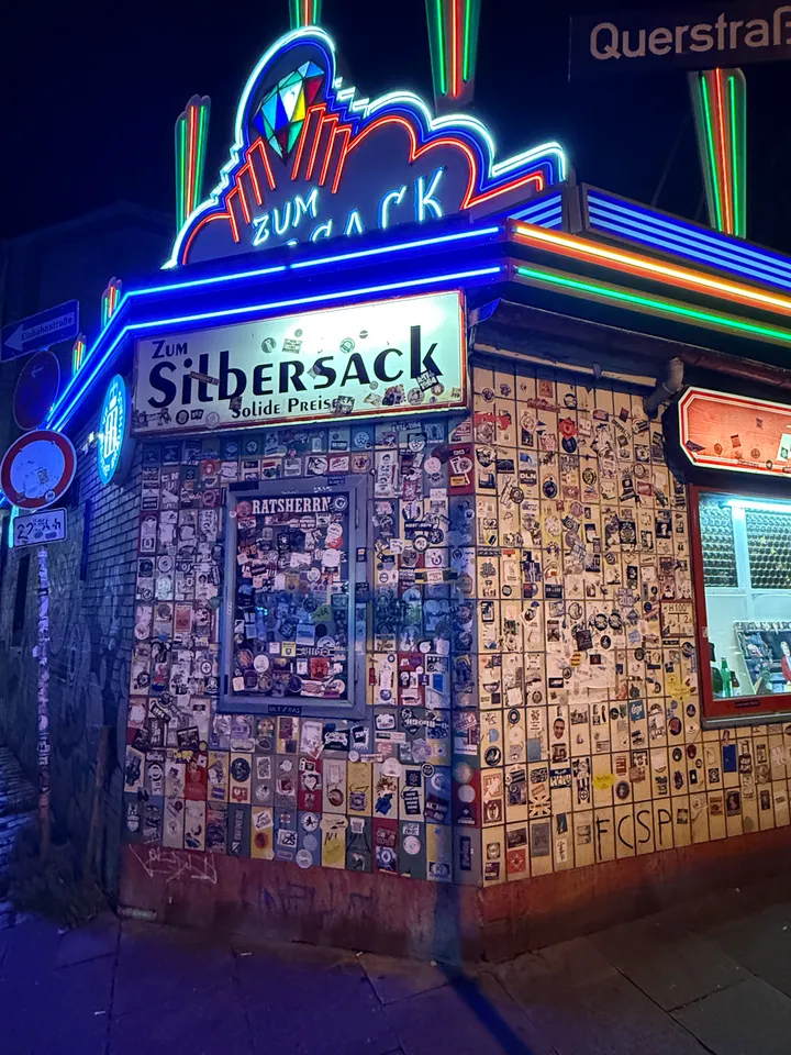 Gekachelte Gebäudeecke voller Aufkleber unter Neonröhren und dem Schild „Zum Silbersack – Solide Preise“