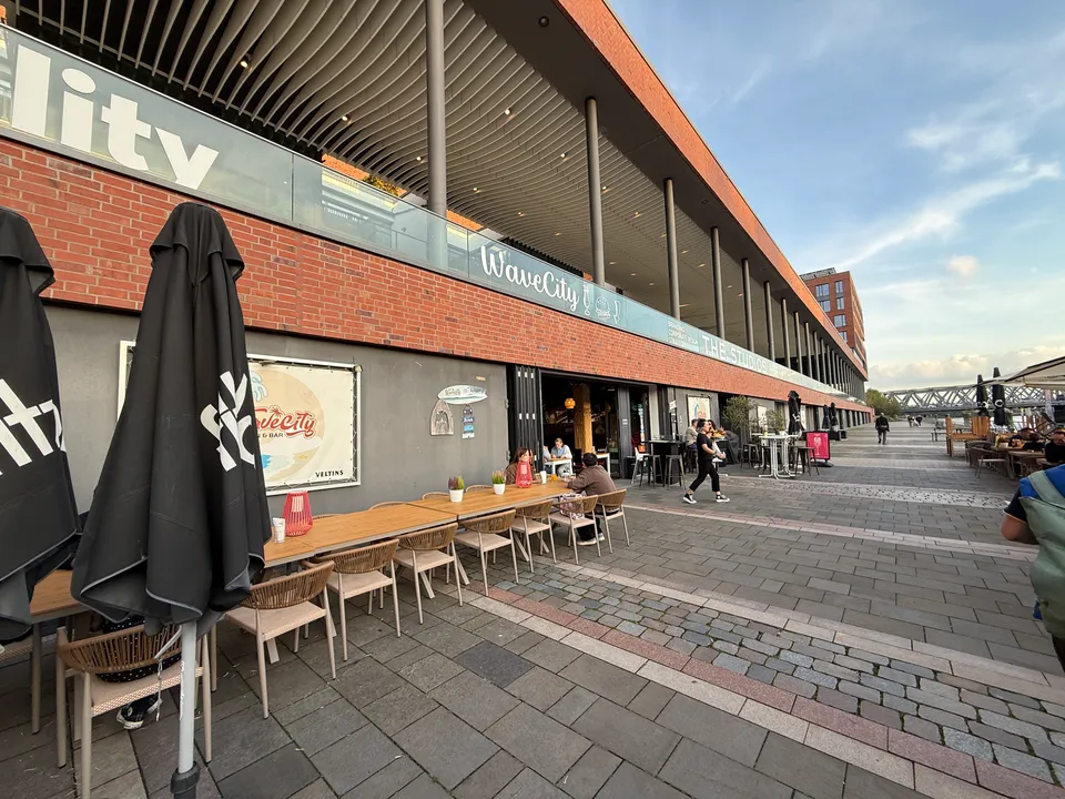 Außenterrasse des Restaurants WaveCity mit Holztischen und Schirmen vor einer Klinkerfassade an der Uferpromenade