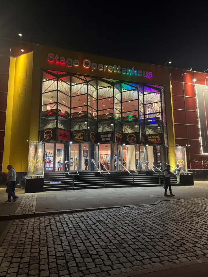Beleuchteter gläserner Eingang des Stage Operettenhauses bei Nacht mit Treppe und Schriftzug in Regenbogenfarben