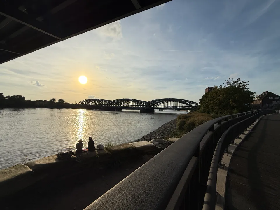 Stahlbogenbrücke über der Elbe im Gegenlicht der tiefen Sonne, vorne Uferweg mit Geländer und zwei sitzende Personen