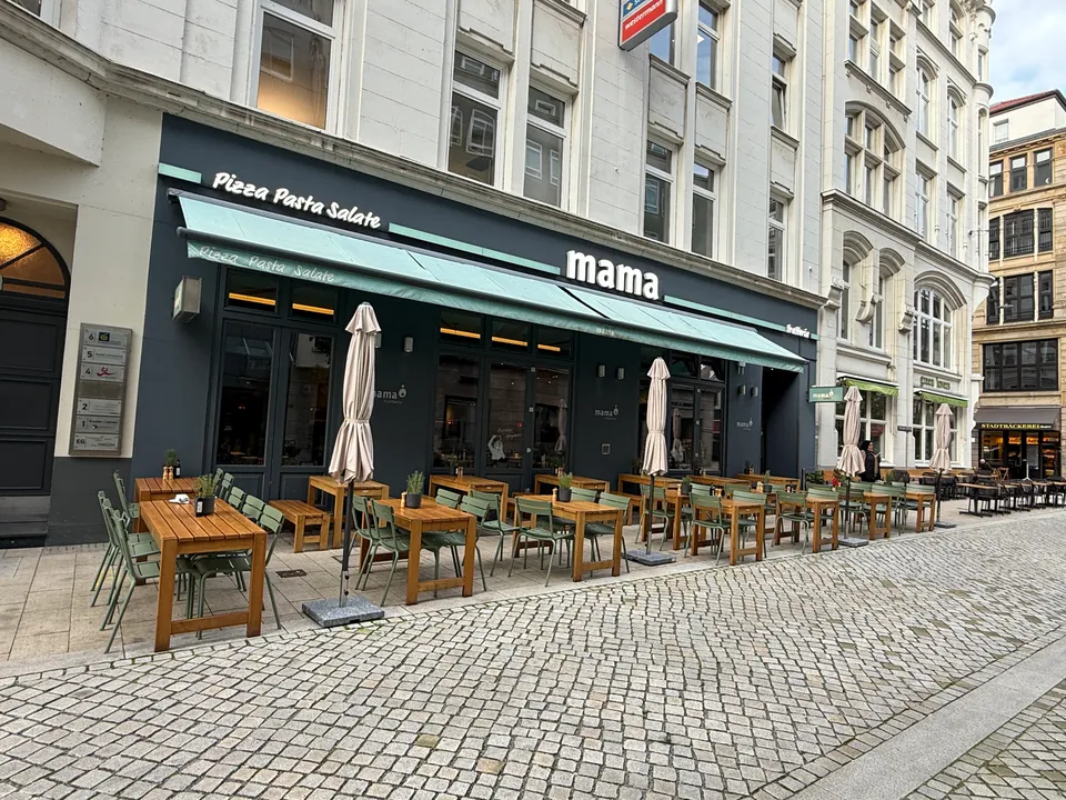 Restaurant mama mit mintgrüner Markise und leeren Holztischen auf dem Gehweg einer Gasse mit Kopfsteinpflaster