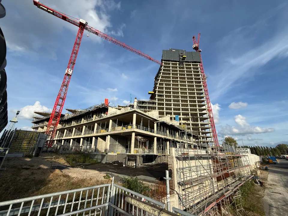 Rohbau eines Hochhauses mit breitem Sockelgebäude, zwei roten Turmdrehkränen, Gerüsten und Bauzaun im Vordergrund