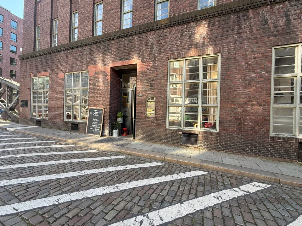 Backsteinfassade mit Café-Eingang, Kreidetafel und Sprossenfenstern an einer Kopfsteinpflasterstraße, links eine Stahlbrücke