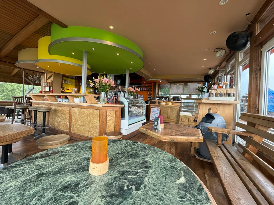 Café-Innenraum mit Holztheke, grünen und gelben Deckenelementen, Kuchenvitrine und Marmortisch im Vordergrund