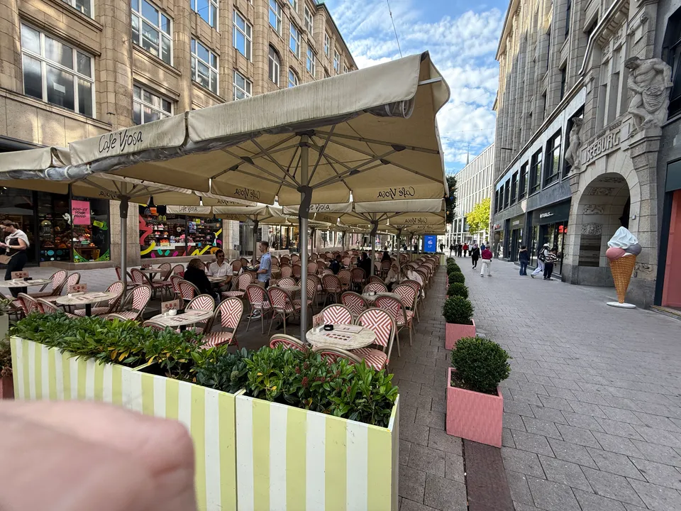 Außenterrasse des Café Vjosa mit beigen Sonnenschirmen, rot-weißen Bistrostühlen und Pflanzkübeln in einer Fußgängerzone