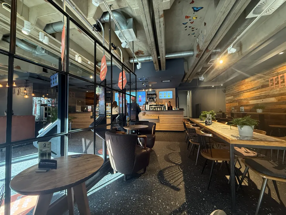 Café-Innenraum mit Fensterfront, runden Holztischen, Ledersesseln, langem Tisch und Theke im Hintergrund