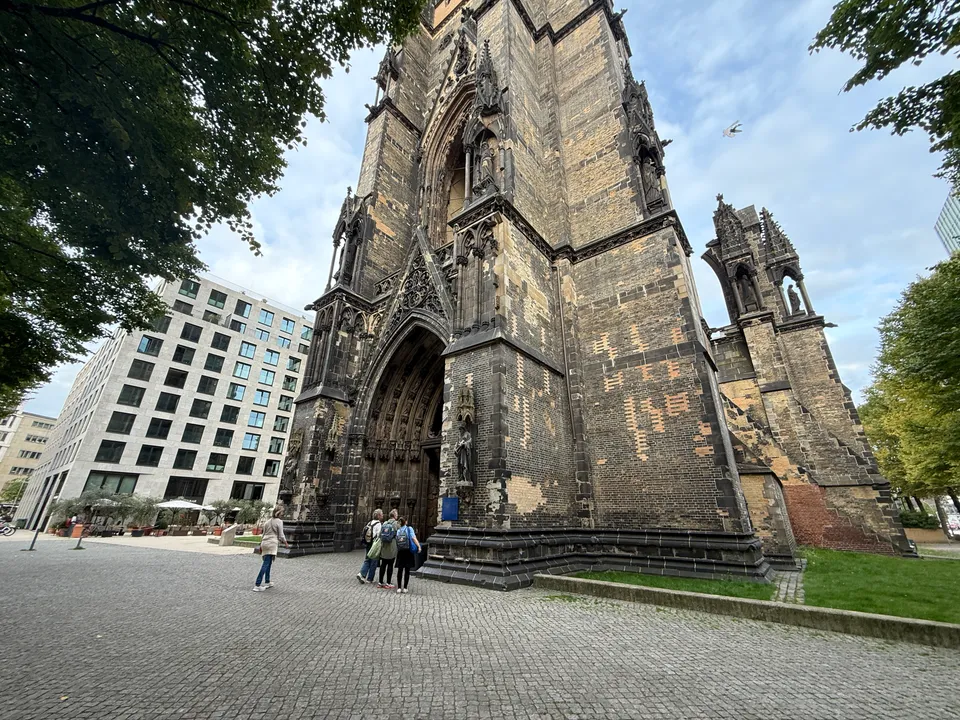 Spitzbogiges Portal am Turm der Kirchenruine St. Nikolai, davor Kopfsteinpflaster, Besuchende und ein modernes Bürogebäude