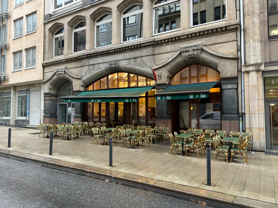 Steinfassade mit Rundbögen, grünen Markisen „Café Paris“ und leeren Bistrotischen und -stühlen auf dem Gehweg