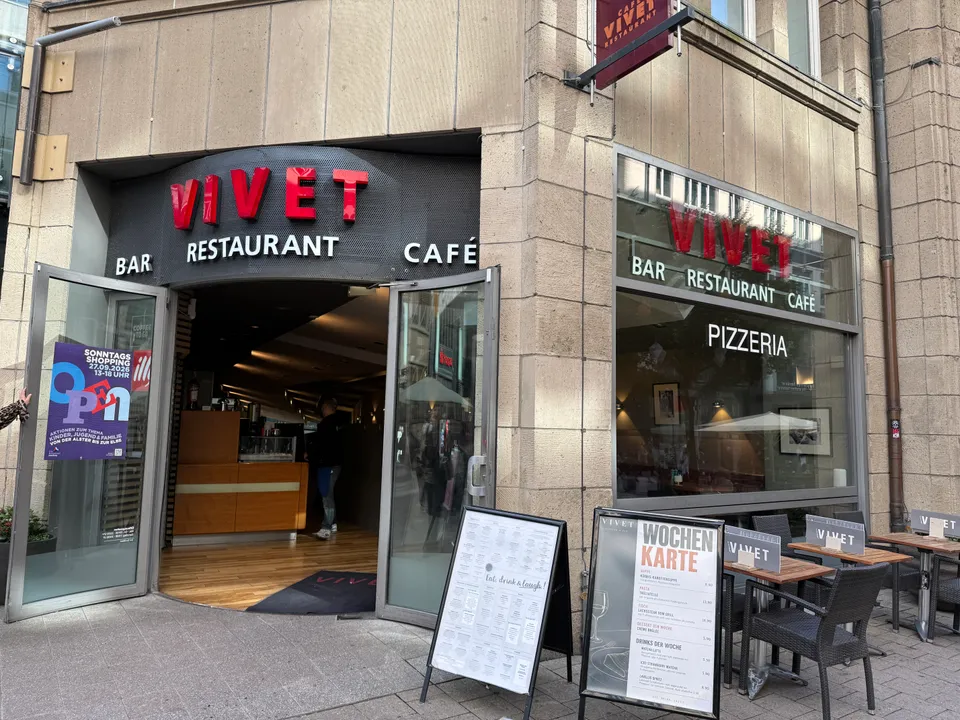 Eingang des Vivet mit roter Leuchtschrift, offenen Glastüren, Menütafeln und Außentischen auf gepflastertem Gehweg