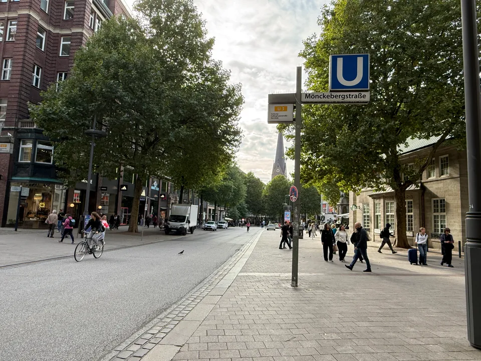 Einkaufsstraße mit U-Bahn-Schild Mönckebergstraße, Fußgängern, Radfahrerin, Bäumen und Kirchturm im Hintergrund