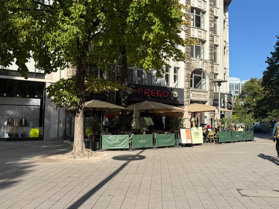 Außenterrasse des Restaurants Prego mit Sonnenschirmen, grünen Bannern und Laubbaum in sonniger Fußgängerzone