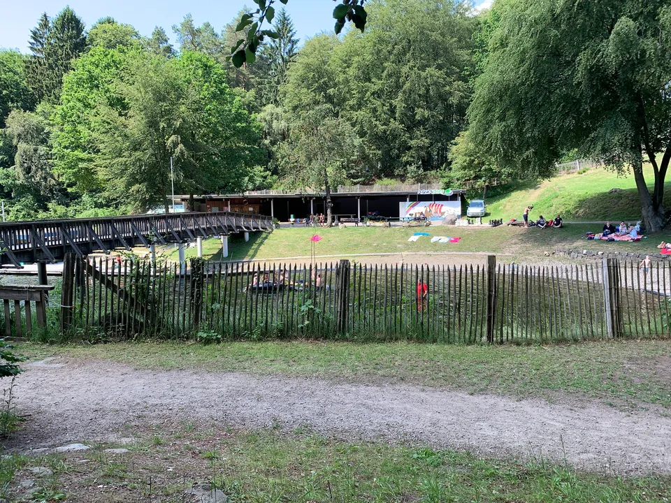 Naturbadesee hinter einem Holzzaun mit Holzbrücke, Liegewiese, flachem Badegebäude und hohen Bäumen bei Sonnenschein