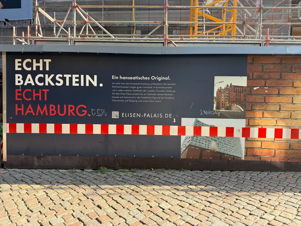 Bauzaun mit Slogan „Echt Backstein. Echt Hamburg.“, rot-weißem Absperrband und Baugerüst dahinter auf Kopfsteinpflaster