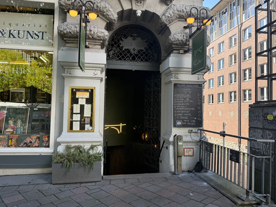 Historischer Rundbogeneingang des Restaurants Rialto mit Schmiedeeisengitter, grünen Hängeschildern und Kreidetafel neben einem Antiquariat