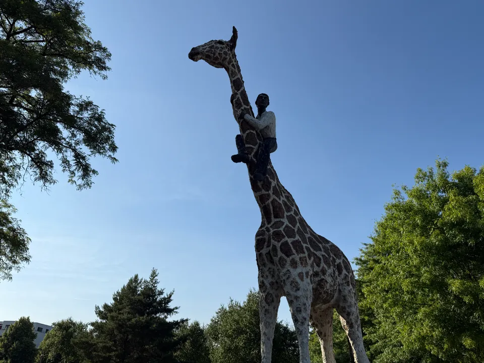Giraffenskulptur mit einer Männerfigur, die sich an den Hals klammert, vor blauem Himmel und Bäumen
