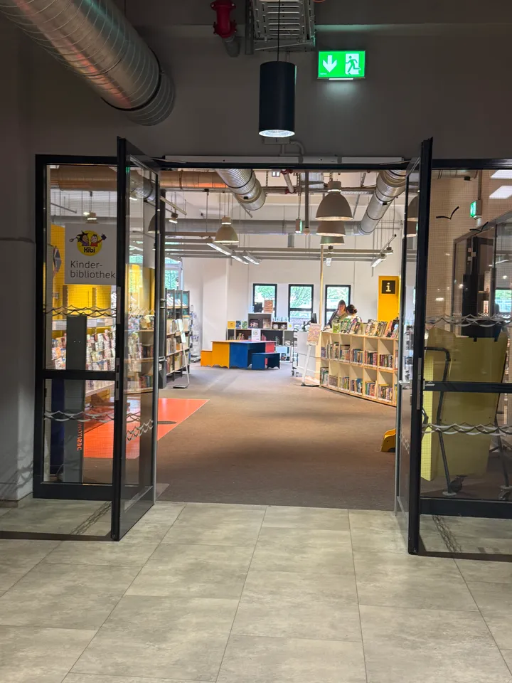 Geöffnete Glastür zur Kinderbibliothek mit Bücherregalen, bunten Sitzmöbeln und Hängeleuchten
