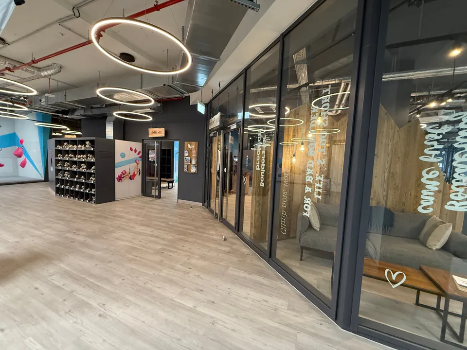 Innenbereich mit Holzboden, Ringleuchten, Regal voller Kletterschuhe und verglaster Lounge mit grauem Sofa
