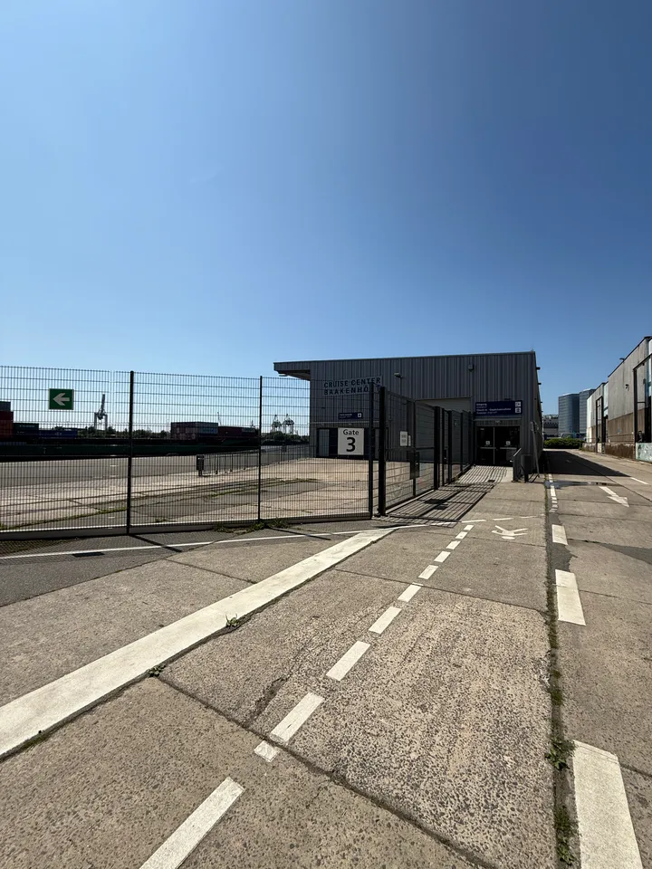 Betonweg entlang eines Gitterzauns mit Schild Gate 3, dahinter die graue Terminalhalle