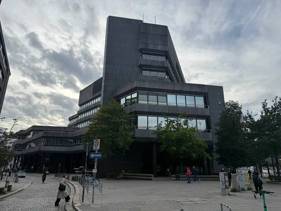Dunkler, gestufter Bürobau mit Fensterbändern hinter Bäumen, davor gepflasterter Platz mit Passanten und Verkehrsschildern