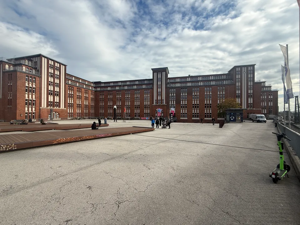Weiter Platz vor großem Backsteingebäude mit Türmen, Sitzpodesten links und E-Scooter rechts, bewölkter Himmel