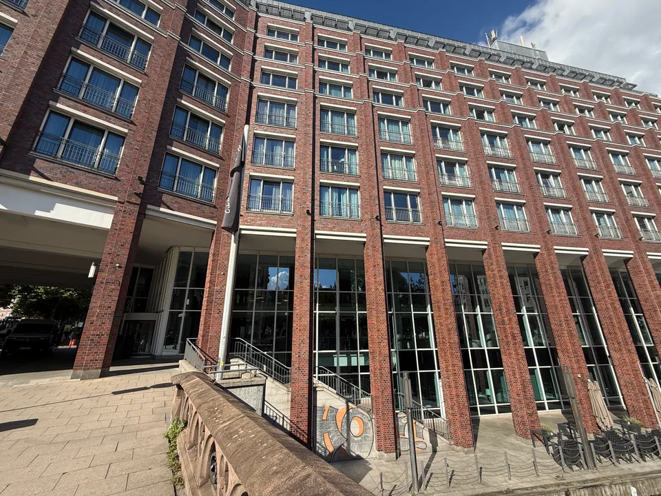 Achtgeschossige Klinkerfassade eines Hotels mit verglastem Erdgeschoss, davor steinerne Brückenbrüstung und Treppe zum Fleet