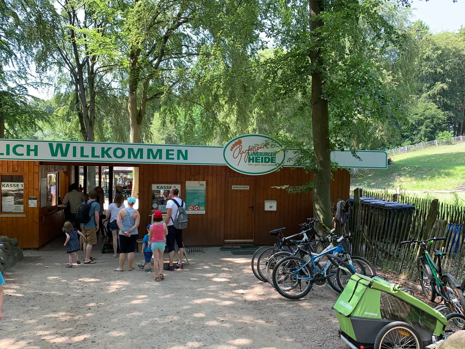 Hölzernes Kassenhaus des Barfußparks mit Willkommensbanner, wartenden Besuchern, abgestellten Fahrrädern und hohen Laubbäumen