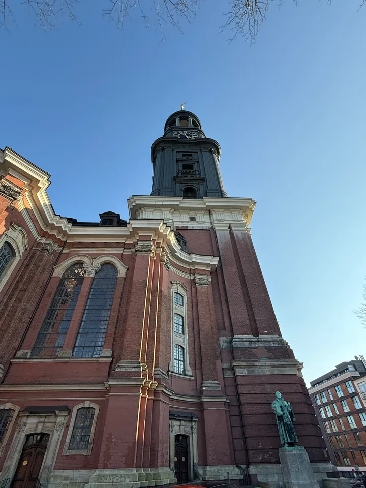 Blick von unten auf den Kirchturm aus Backstein mit Uhr, davor eine Bronzestatue auf Sockel